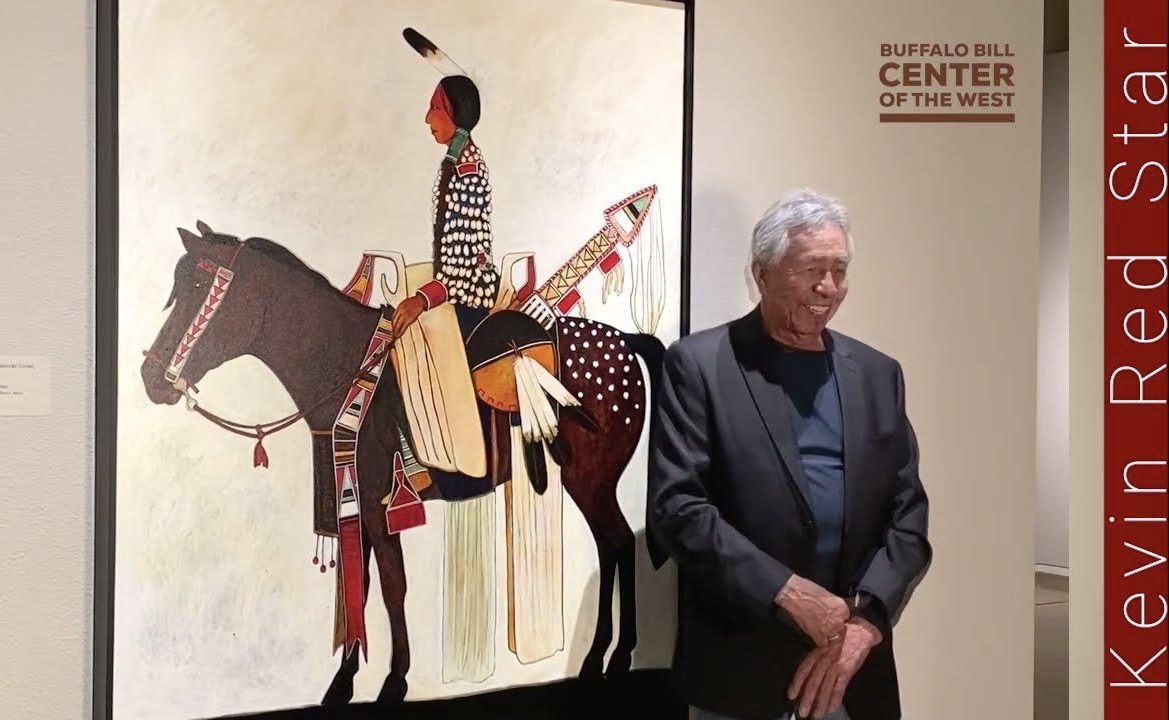 Elderly man stands smiling beside Native American warrior painting at Buffalo Bill Center.